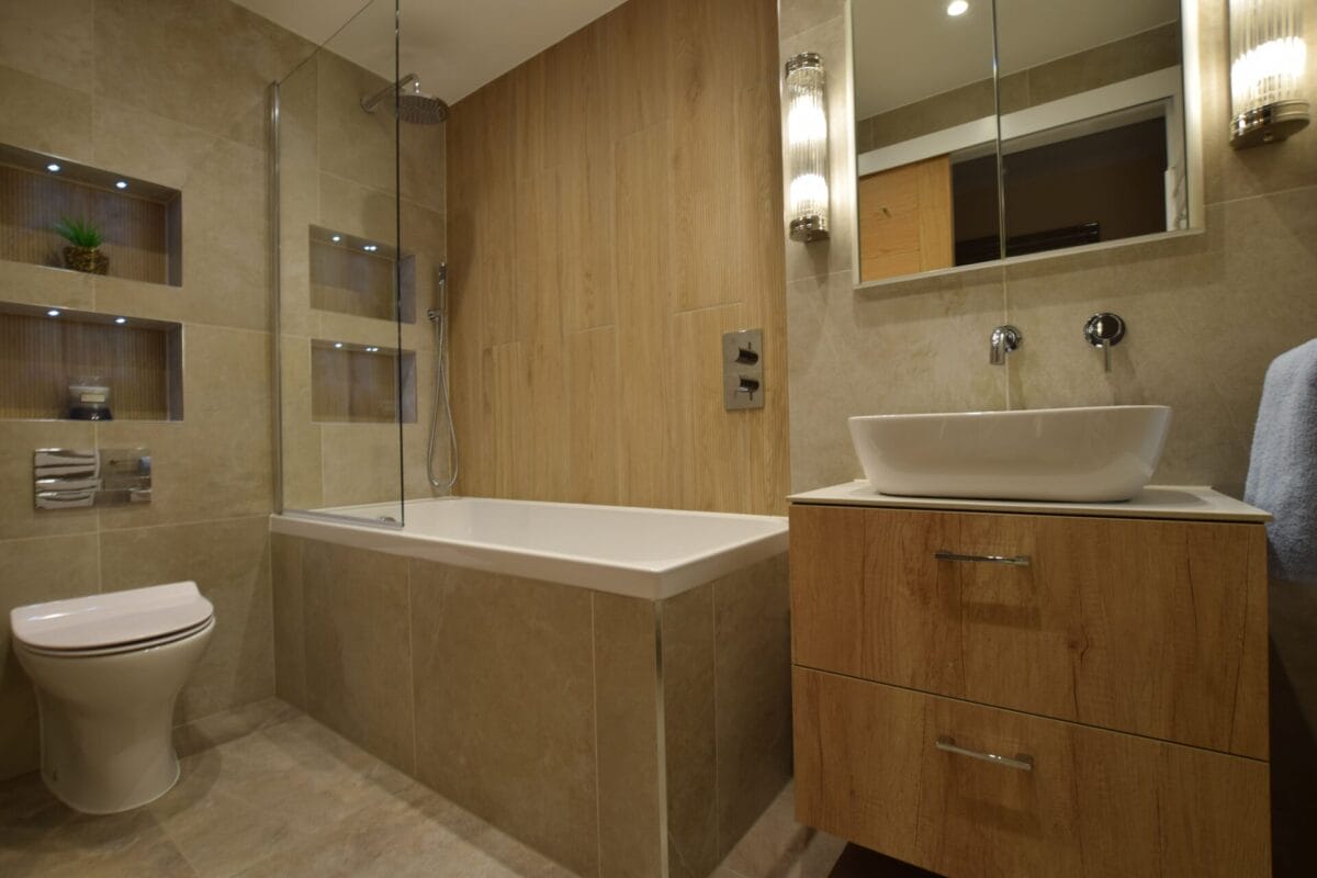 Contemporary family bathroom with wood-effect wall panels, freestanding vessel sink on oak vanity, built-in lit niches and bath with overhead shower — designed and installed by The London Bath Co.