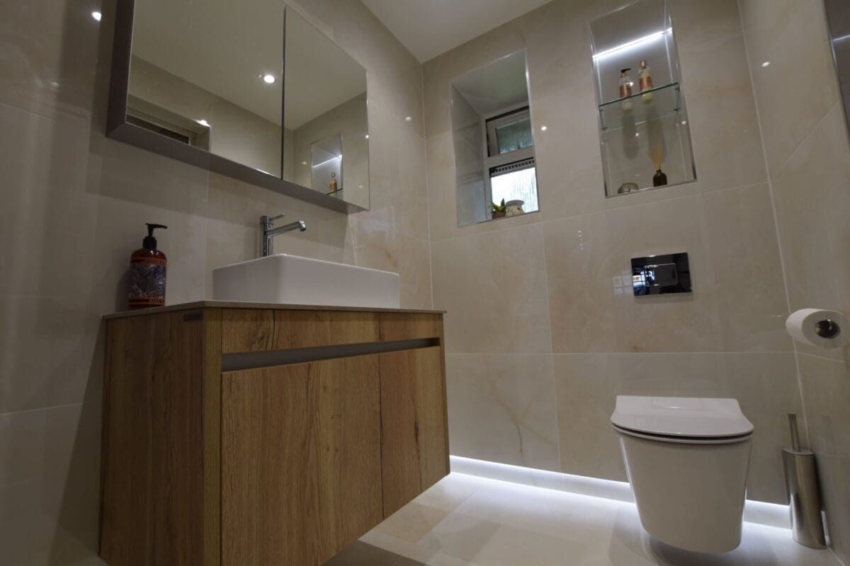 Modern bathroom with large-format cream tiles, wall-hung WC, floating oak vanity unit with vessel basin, LED plinth lighting and recessed shelving — The London Bath Co. London