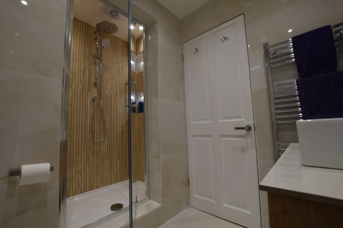 Compact shower enclosure with wood-effect slat tile panel, chrome rainhead and slide rail shower, large-format cream wall tiles and chrome heated towel rail — bathroom installation by The London Bath Co., London