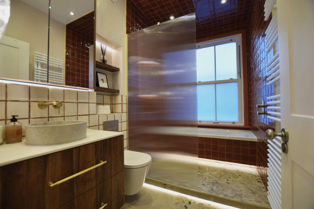 Bold bathroom renovation with deep red gloss square tiles, fluted glass shower screen, terrazzo flooring, stone vessel basin on walnut vanity with brass fixtures and LED plinth lighting — The London Bath Co., London
