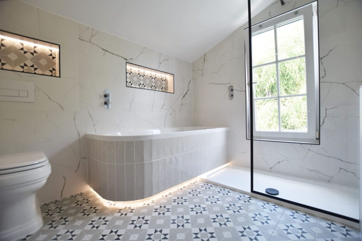 Period property bathroom with Calacatta marble-effect wall tiles, monochrome patterned encaustic floor tiles, curved corner bath with vertical tile panel and under-bath LED strip lighting — The London Bath Co.