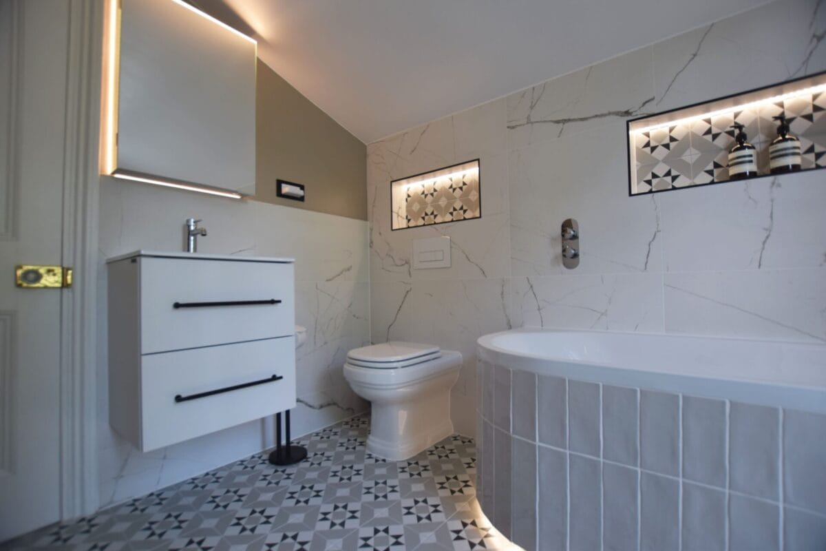Full bathroom renovation in a London period home with white floating vanity unit, black hardware, Calacatta marble-effect tiles, lit recessed niches with patterned tile inserts and corner bath — The London Bath Co.